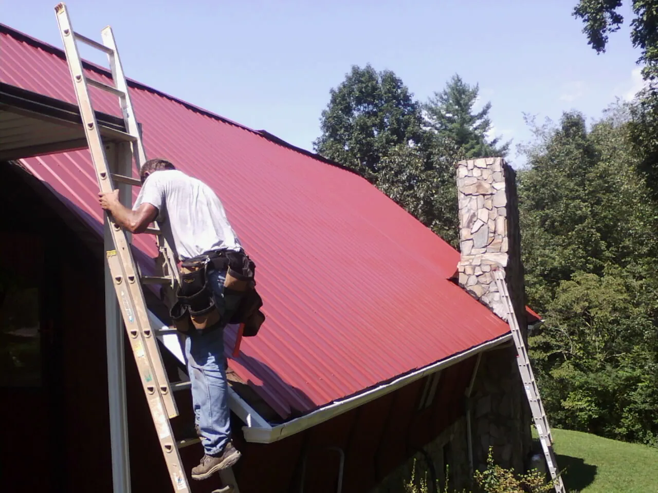 Roofer on a ladder beside a red metal roof and stone chimney — Soffit & Fascia Repair by Roofing Brothers in East Wareham